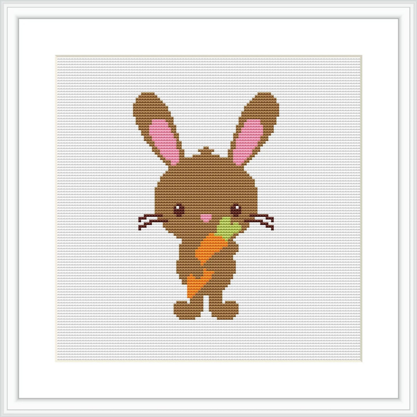 The image shows a framed cross stitch pattern of a cute brown bunny with pink inner ears standing upright. The bunny is holding a carrot with green leaves in its front paws. The background is white, and the stitching is visible.