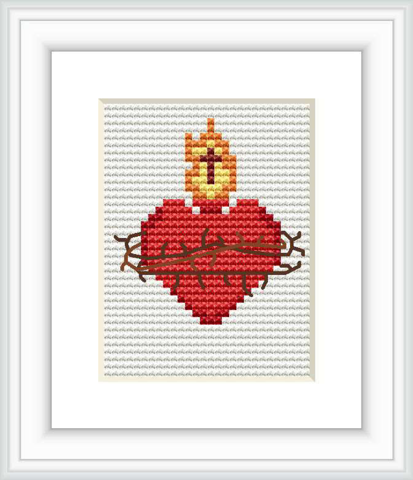 The image showcases a cross stitch pattern framed and displayed on a wall. The pattern features a stylized red heart with a yellow and orange flame on top, all outlined by brown thorny branches, on a white stitched background.