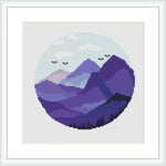 The image features a round cross-stitch pattern showing a stylized mountain landscape within a circular frame. The mountains are portrayed in various shades of purple and are complemented by a pale sky with small birds flying.