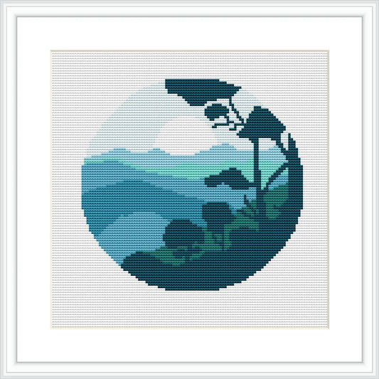 The cross stitch pattern depicts a circular landscape scene with a range of mountains in the background. It includes various shades of green for the mountains and lighter shades for the sky. A simplistic tree figure appears in the foreground on the right.