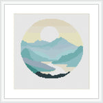 The cross stitch pattern depicts a circular, framed landscape scene featuring a river, rolling mountains in layered hues, and a stylized sun peeking from behind the mountain peaks. The river is in the foreground and the whole scene is set within a round border.