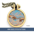 The image shows a small cross stitch pendant featuring two outstretched hands reaching towards each other against a sky-like background, referencing 'The Creation of Adam'. The pendant is encased in a circular frame with a chain attached.