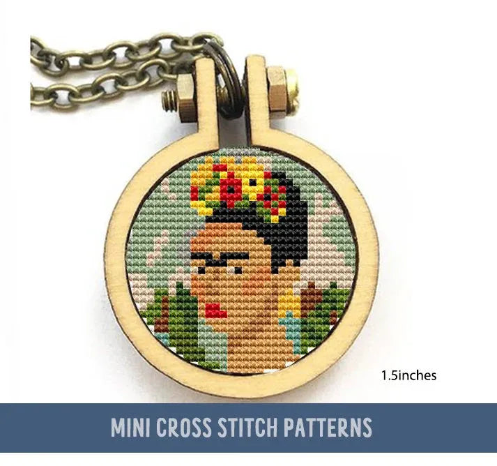 The cross stitch features a stylized portrait reminiscent of Frida Kahlo, with floral elements in her hair. The design lies within a circular frame with a bronze chain attached.