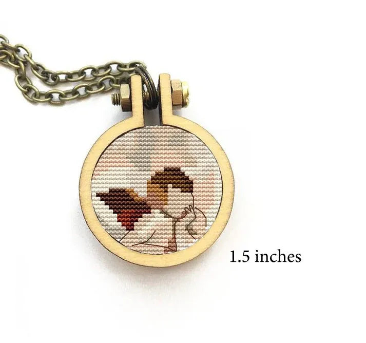 The image features a small cross stitch pattern in a round pendant showing a cherub from Raphael's famous painting. The pendant is attached to a chain.