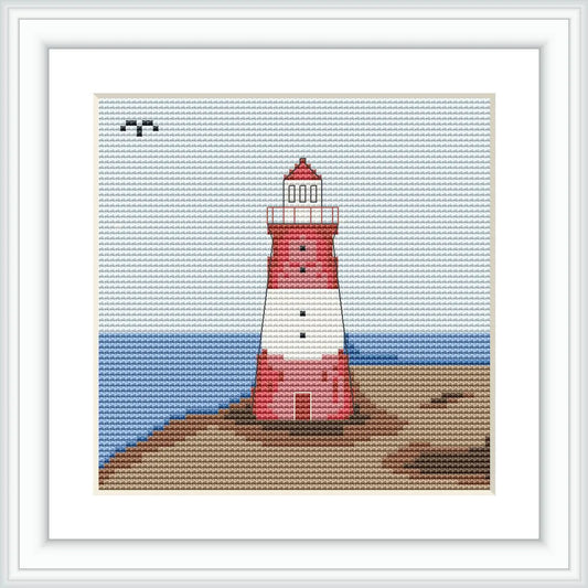 The image shows a framed cross stitch pattern of a red and white lighthouse set against a clear sky and blue sea backdrop, with a sandy foreground.