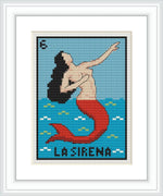 The image displays a framed cross stitch pattern of 'La Sirena', a figure from the traditional Mexican game 'La Loteria'. The pattern shows a mermaid with a red tail and black hair, posed against a blue backdrop that represents the sea, with lighter blue pixelated waves near the bottom.