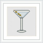 The image depicts a cross-stitch pattern of a martini glass filled with a drink and garnished with three olives on a toothpick. The glass is centrally placed within a white-background frame.