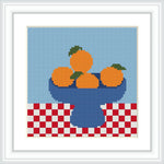 The image shows a blue bowl with four oranges, each with a small green leaf. The bowl sits on a red and white checkerboard tablecloth with a blue background.