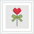 The image depicts a cross stitch pattern of a red heart-shaped lollipop tied with a green bow on a white background.