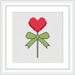 The image depicts a cross stitch pattern of a red heart-shaped lollipop tied with a green bow on a white background.