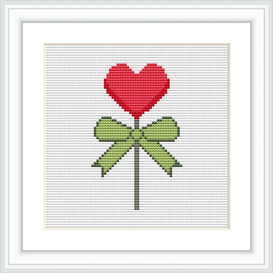 The image depicts a cross stitch pattern of a red heart-shaped lollipop tied with a green bow on a white background.