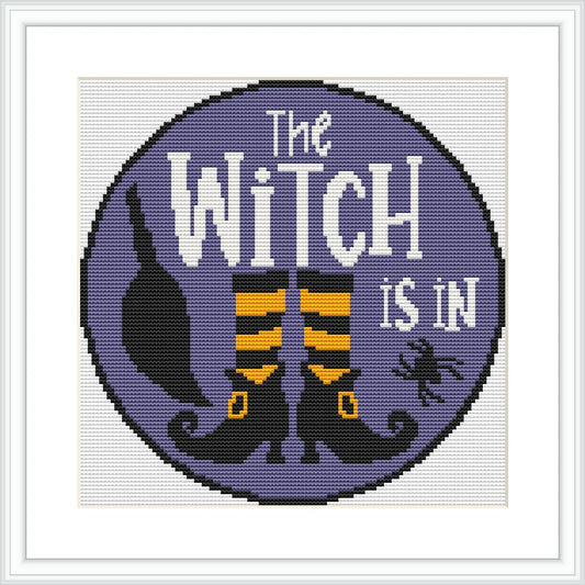 The cross stitch design features a circular frame filled with a purple background. In the foreground, there is a witch's hat, striped stockings with witch shoes, and a small spider to the right. The words 'The Witch is In' are prominently displayed.