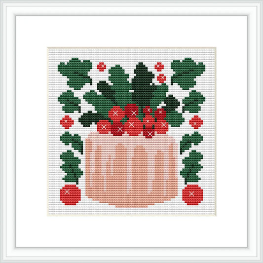 The cross stitch pattern depicts a pink cake topped with green leaves and red berries. The cake is placed centrally within a square frame, surrounded by symmetric arrangements of leaves and berries.