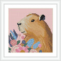 A capybara is depicted in a cross stitch design, placed centrally and surrounded by a variety of colorful flowers on a pink background.