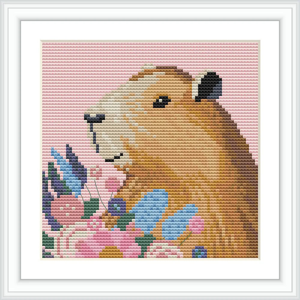A capybara is depicted in a cross stitch design, placed centrally and surrounded by a variety of colorful flowers on a pink background.
