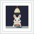 A white bunny with pink ears sitting below a hanging light. The bunny is holding a blue bowl filled with orange carrots on a light-colored surface.