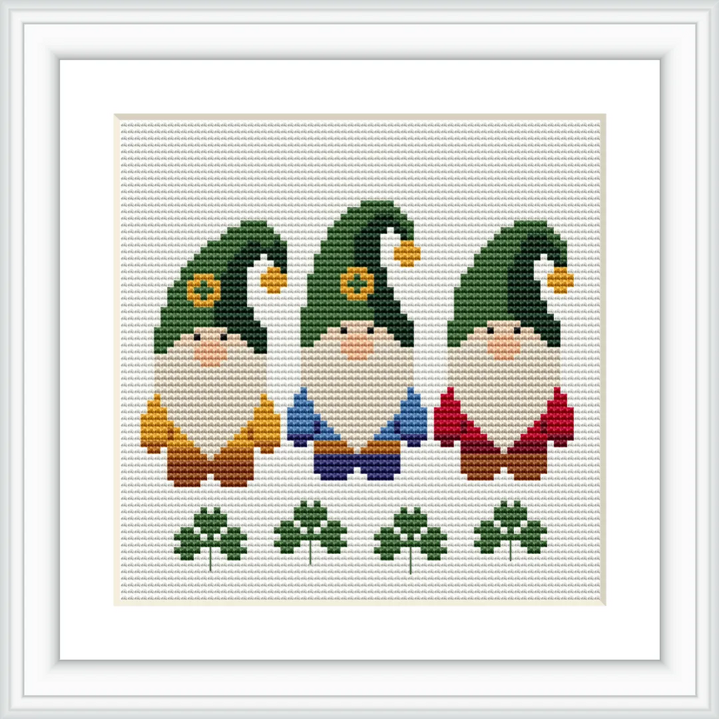 The image features three gnomes standing in a row, each with a green hat and unique colored clothing—yellow, blue, and red. Below them, there are three green clovers. The background is a plain white cross-stitched fabric.