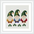 The image features three gnomes standing in a row, each with a green hat and unique colored clothing—yellow, blue, and red. Below them, there are three green clovers. The background is a plain white cross-stitched fabric.