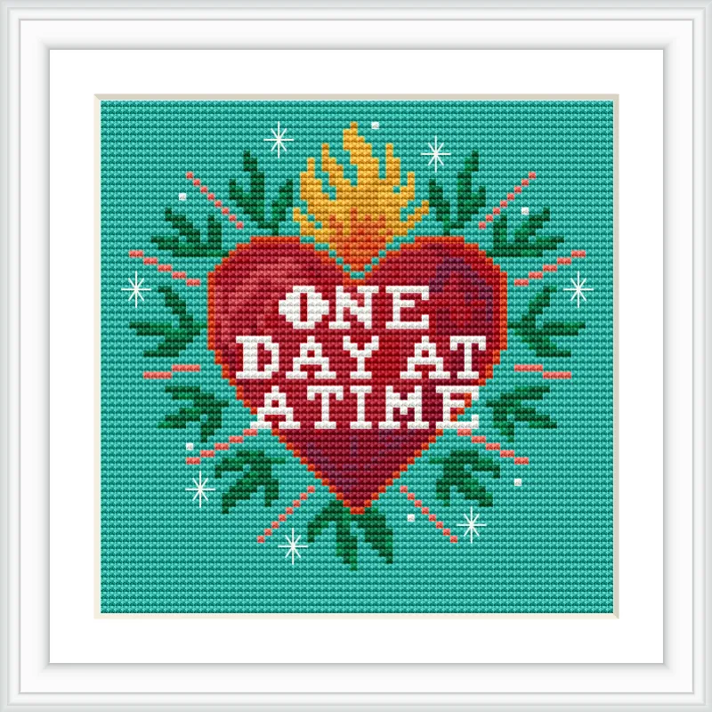 The image features a cross stitch pattern with a red heart in the center, surrounded by green leaves and a yellow flame on top. The heart contains the words 'One Day at a Time' in white text, set against a teal background with decorative star-like accents.