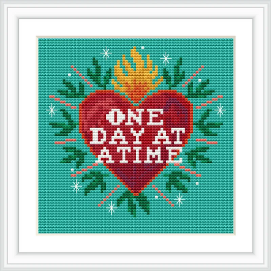 The image features a cross stitch pattern with a red heart in the center, surrounded by green leaves and a yellow flame on top. The heart contains the words 'One Day at a Time' in white text, set against a teal background with decorative star-like accents.