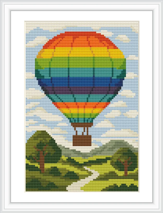 The image shows a large, rainbow-colored hot air balloon floating above a green landscape with a winding path and trees under a blue sky scattered with clouds.