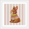 A cute plush bunny bear is sitting upright, wearing a colorful striped sweater against a background of vertical pink and white stripes.