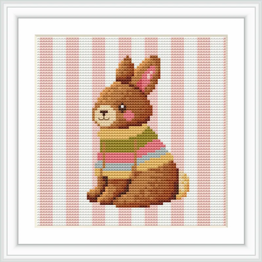 A cute plush bunny bear is sitting upright, wearing a colorful striped sweater against a background of vertical pink and white stripes.