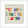 The image features a framed cross stitch with the text 'This Room is Made for Playing' in multicolored letters, bordered by a light blue scalloped edge.
