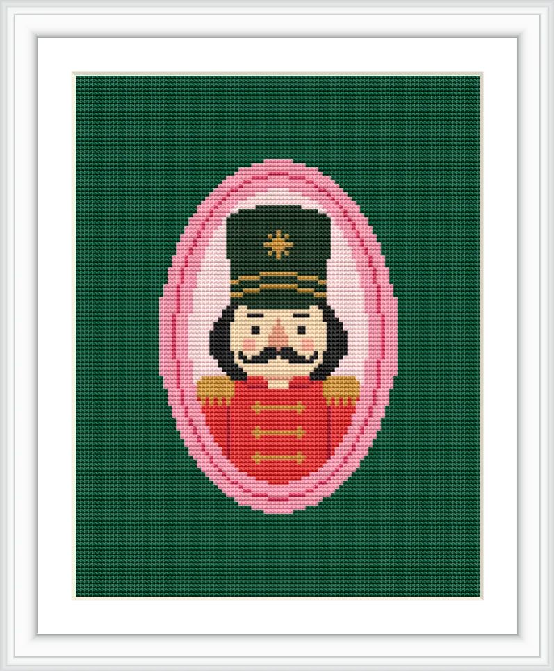 The image features a portrait of a nutcracker soldier framed in a pink oval against a dark green background. The soldier is dressed in red with gold details and wears a black hat.