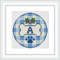 The image shows a cross stitch design featuring a blue checkerboard background with a central monogram letter 'A'. Above the letter, there's a holly motif with green leaves and red berries. The design is enclosed within a circular border.