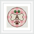 Cross stitch pattern features a circular design with a bone in the center, bordered by a pink checkered pattern. Above the bone is a floral decoration, and below is a paw print. The letter 'A' is prominently displayed on the bone.