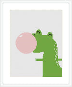 The image displays a framed cross stitch pattern depicting a stylized green dinosaur facing left, blowing a large pink bubble from its mouth. The background is plain white, highlighting the dinosaur and the bubble.