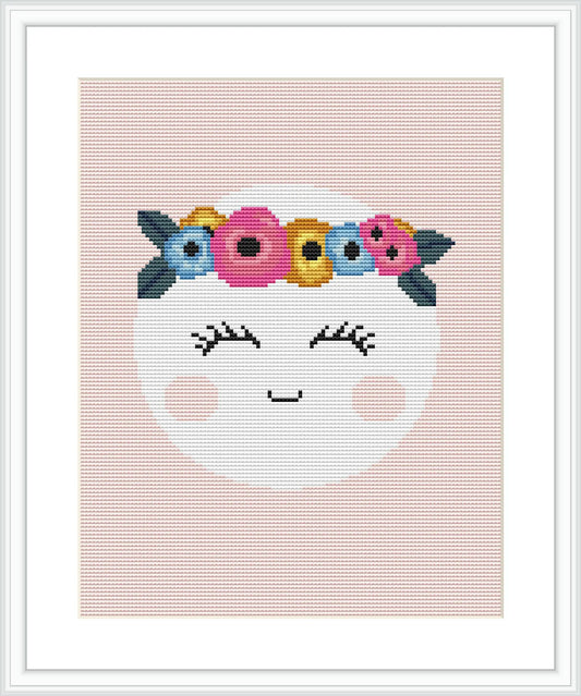 The image features a cross stitch pattern design portraying a smiling crescent moon with closed eyes and lush cheeks. The moon is crowned with a floral headband composed of various colorful flowers. The design is displayed on a pink background and framed with a white border.