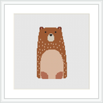 The image presents a cross stitch pattern depicting a stylized bear. The bear is designed with various shades of brown and has a large beige section on its stomach and snout. Its eyes and nose are stitched in black, standing out against the lighter shades. The embroidery is shown as if it's been framed on a wall.