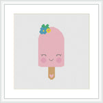 The image depicts a framed cross stitch pattern of a pink popsicle with a smiling face on the popsicle's body. The popsicle stick is brown, and on top of the popsicle, there are what appear to be blueberry and mint leaf embellishments. The design is set against a white aida cloth background, held within a white frame.