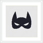 The image displays a black superhero mask silhouette centered on a white cross stitch canvas, framed by a simple light frame and matted with white, providing stark contrast.