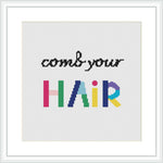 The image depicts a cross stitch pattern with the phrase 'comb your HAIR', where 'HAIR' is written in large, colorful block letters with each character displaying a different color of the rainbow. The rest of the phrase is in smaller black letters. The design is centered on a white aida cloth within a simple, square frame.