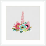 The cross stitch pattern displays a whimsical unicorn horn in shades of pink with a twist, surrounded by an array of colorful flowers and green leaves, framed in a simple white border.