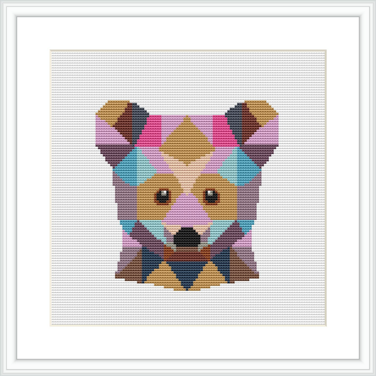 The image shows a framed cross stitch pattern of a bear's face, composed of various geometric shapes in a range of pastel colors. The pattern is centered and showcased within a square frame against a white background.