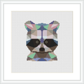 The image depicts a framed cross stitch piece featuring a stylized raccoon face comprised of geometric shapes. The raccoon is centered on a white fabric background and is rendered in a variety of colors including earth tones and vibrant accents.