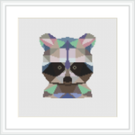 The image depicts a framed cross stitch piece featuring a stylized raccoon face comprised of geometric shapes. The raccoon is centered on a white fabric background and is rendered in a variety of colors including earth tones and vibrant accents.