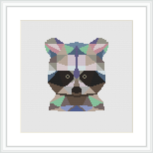 The image depicts a framed cross stitch piece featuring a stylized raccoon face comprised of geometric shapes. The raccoon is centered on a white fabric background and is rendered in a variety of colors including earth tones and vibrant accents.