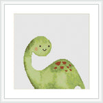 The image displays a framed cross stitch pattern featuring a cartoon-style green dinosaur with red spots on its back, smiling against a white background.