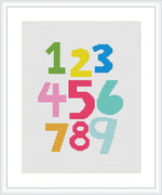 The image shows a cross stitch pattern featuring numbers 1 through 9, each embroidered in a different color, on a white grid-like fabric. The numbers are in bold fonts and are arranged in three rows of three.