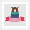 The image shows a cross stitch design featuring a brown bear wearing a pink Santa hat and partially wrapped in a gift box with layers of green, blue, and purple. The words 'MERRY CHRISTMAS' are stitched in red on a banner below the bear.