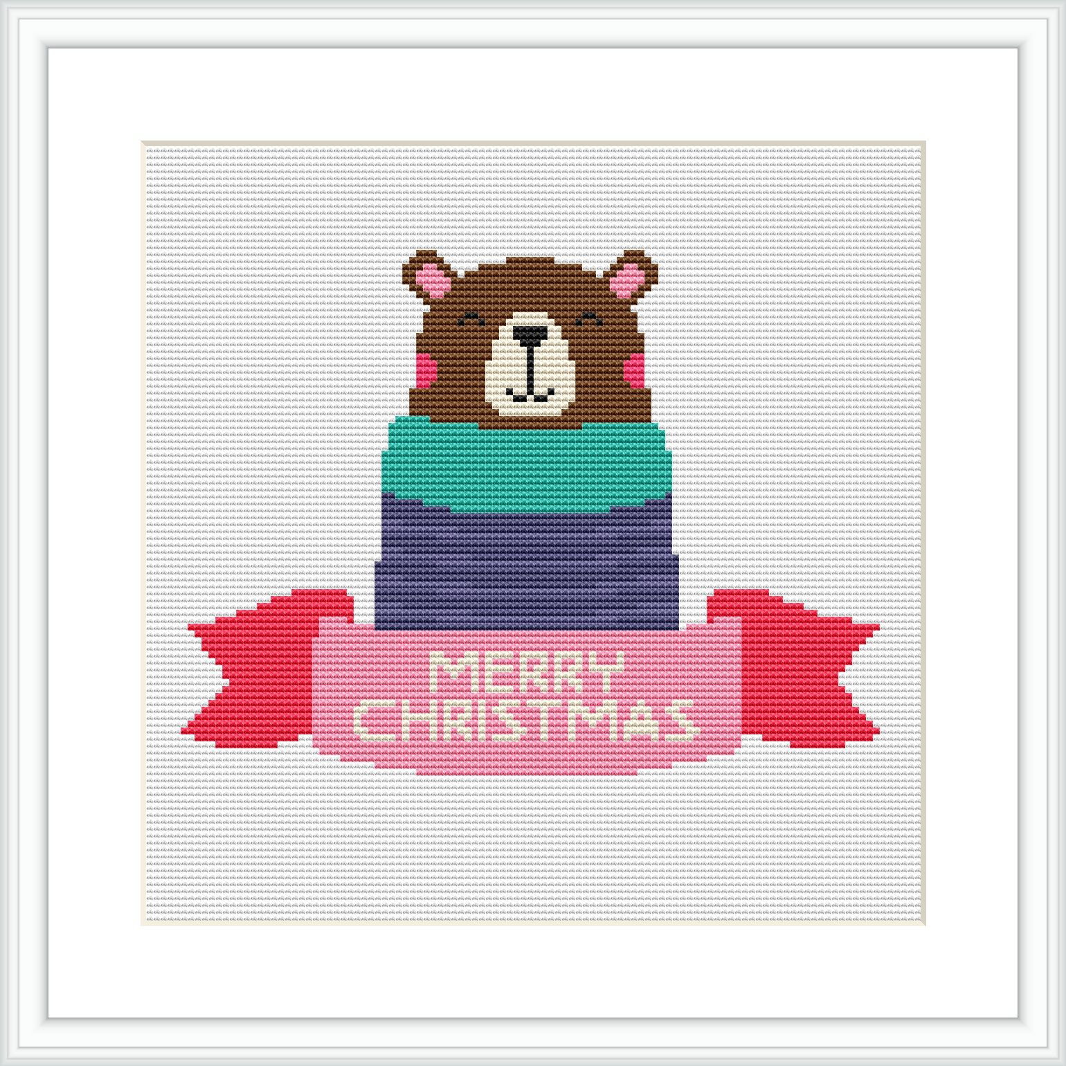 The image shows a cross stitch design featuring a brown bear wearing a pink Santa hat and partially wrapped in a gift box with layers of green, blue, and purple. The words 'MERRY CHRISTMAS' are stitched in red on a banner below the bear.