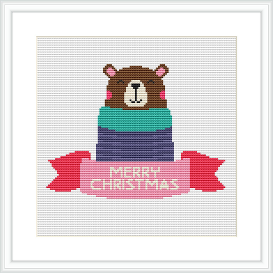 The image shows a cross stitch design featuring a brown bear wearing a pink Santa hat and partially wrapped in a gift box with layers of green, blue, and purple. The words 'MERRY CHRISTMAS' are stitched in red on a banner below the bear.