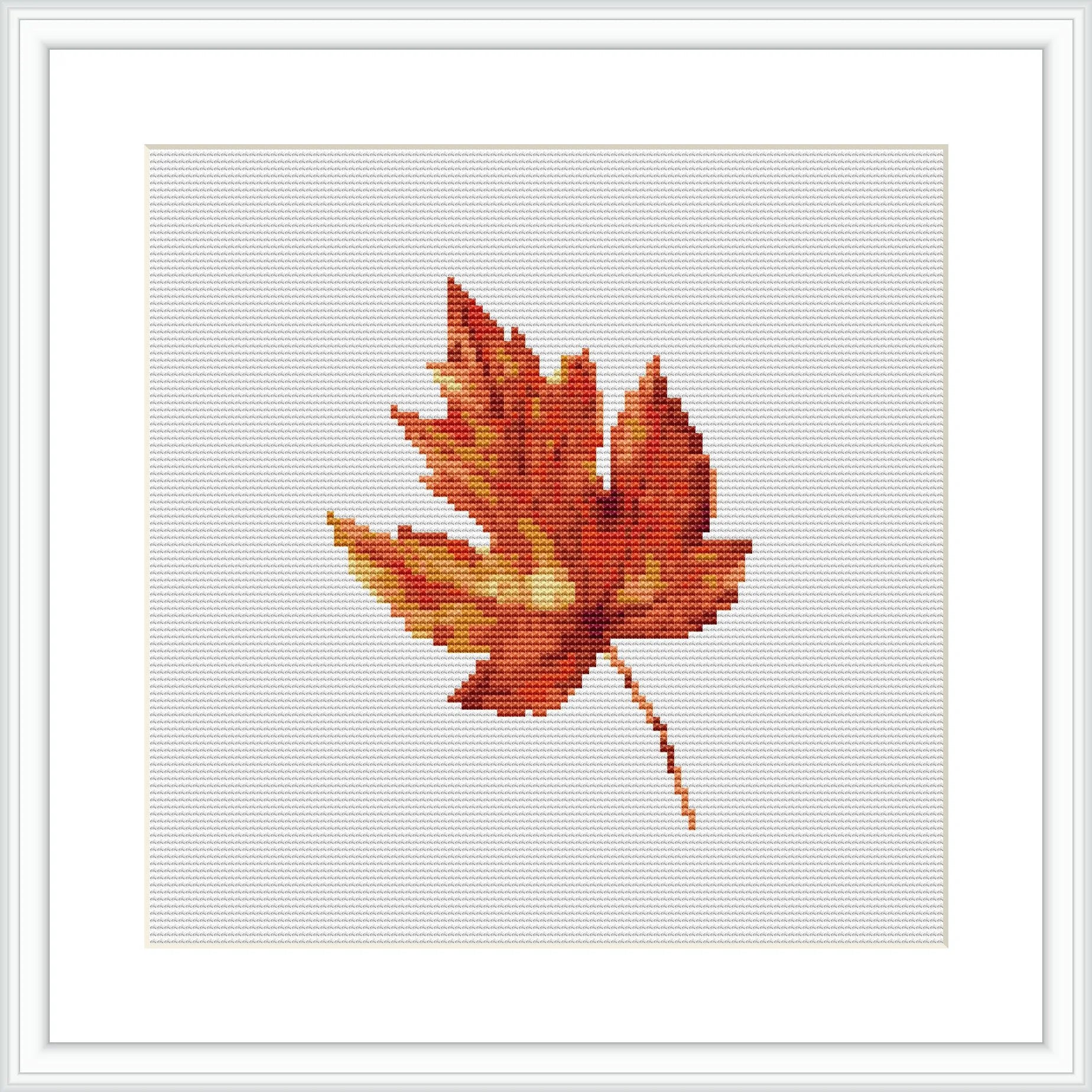 The image displays a single autumn leaf with a spectrum of warm colors, intricately designed using cross stitch techniques. The leaf is centered on a white cloth and has variations of brown, orange, and yellow hues, encased in a simple white frame.