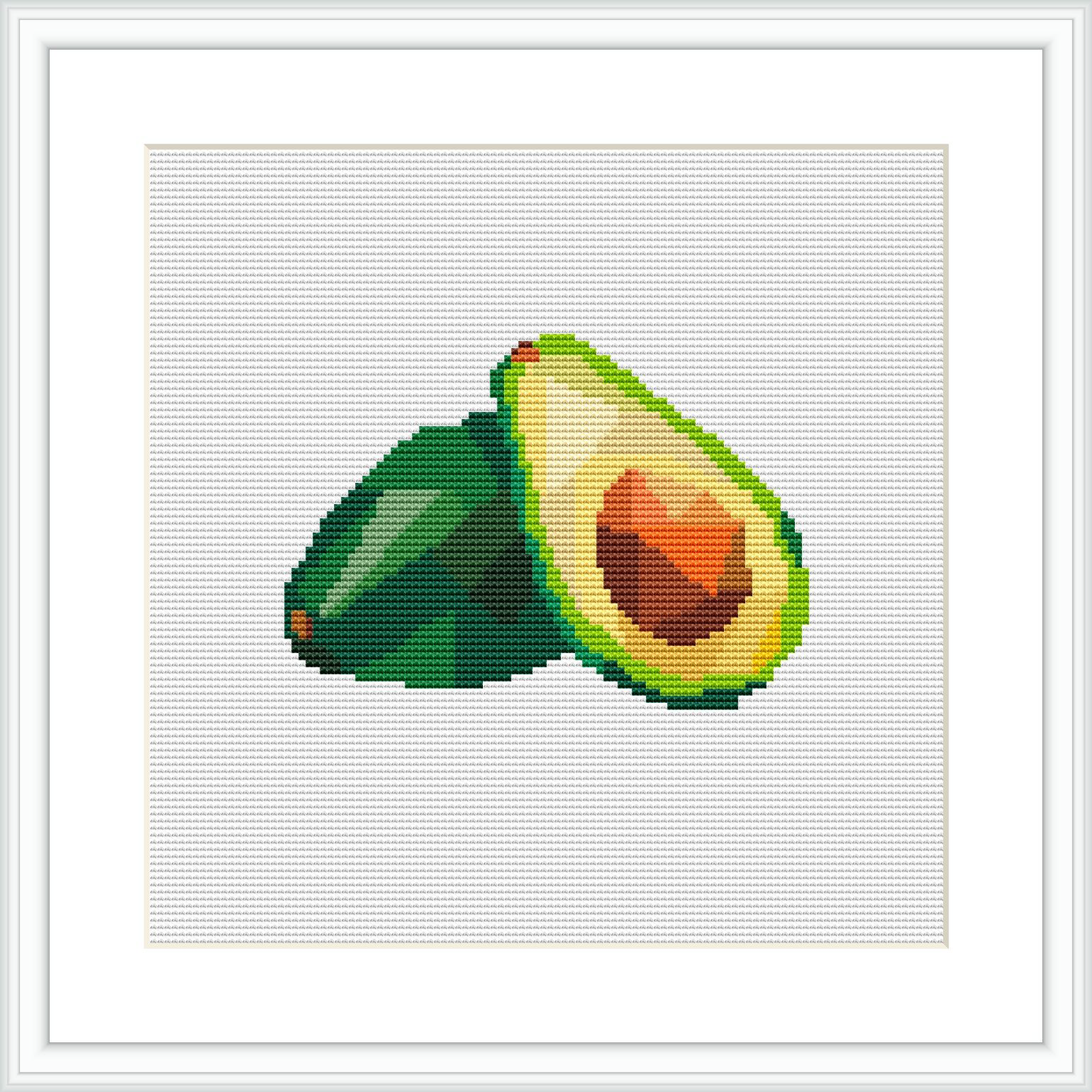 The image shows a cross stitch pattern of an avocado cut in half with the pit visible. One half of the avocado is resting with the flat side down, while the other half stands upright, showcasing the inside. The stitch work is visible against a white canvas, and the design is framed by a simple square frame.