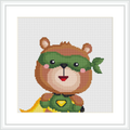 The image displays a cute, cartoon-style bear wearing a green mask and a hero’s cape. The bear also has a green emblem on its chest, all crafted using cross stitch technique on a white background.